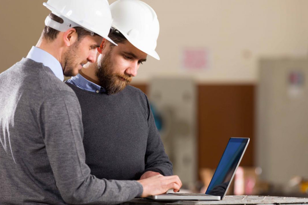Two men dressed in smart casual with construction hard hats looking at data on a laptop computer within and industrial setting 
