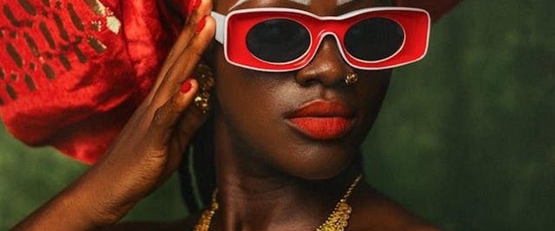 A person wearing red-framed sunglasses and a bold red headwrap poses confidently. They have vibrant red lipstick and ornate gold jewelry, set against a green backdrop.